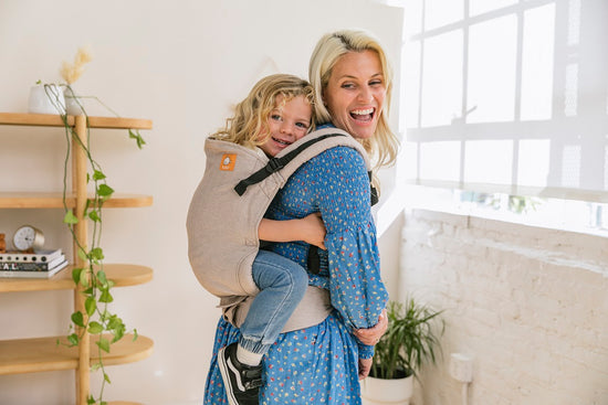 Happy mum carrying her toddler in back carry position in Tula Linen Toddler Carrier