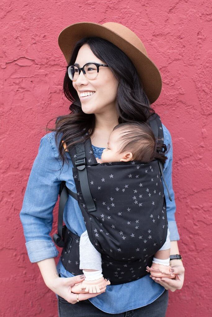 Newborn being worn in a hip healthy carrier suitable from birth with a print of gray star clusters cascading across a black background