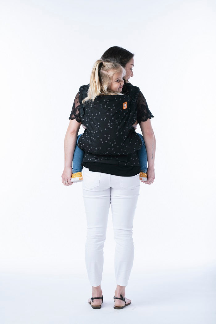 A mother carrying her daughter in back-carry position in the Toddler Carrier Discover from Tula.