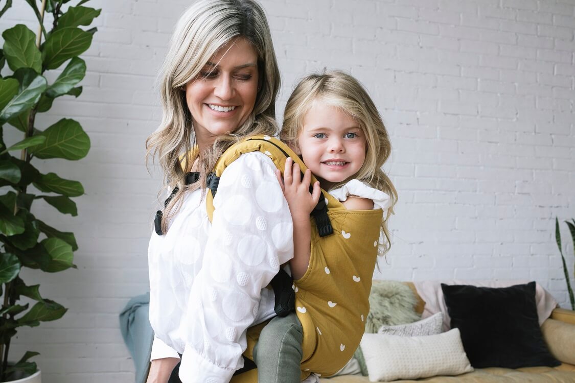 Mum wearing a preschool girl in a back carry in an ergomic preschool carrier by Baby Tula.