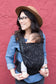Newborn being worn in a hip healthy carrier suitable from birth with a print of gray star clusters cascading across a black background