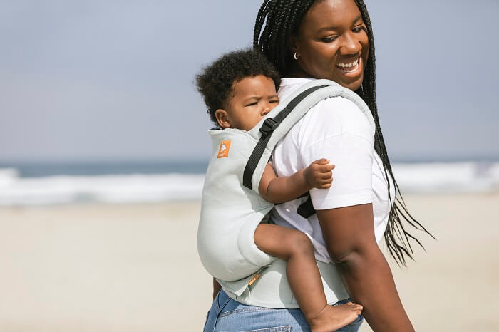 A woman at the beach carrying a smiling baby sitting in the ergonomic Free-to-Grow Linen Baby Carrier Seafoam in the front-carry position.