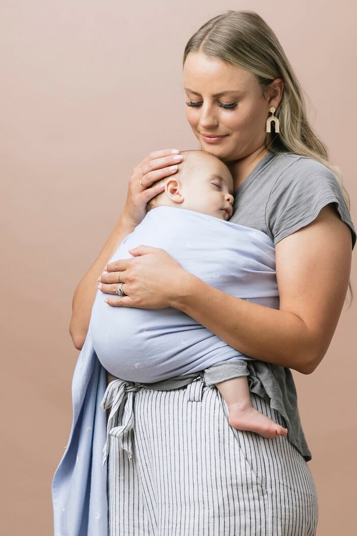 Mum wearing sleeping newborn in Tula ring sling carrier from woven fabric in powder blue with white moons.