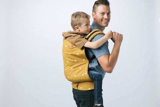 Caregiver wearing a preschooler in a back carry in an ergomic preschool carrier by Baby Tula.