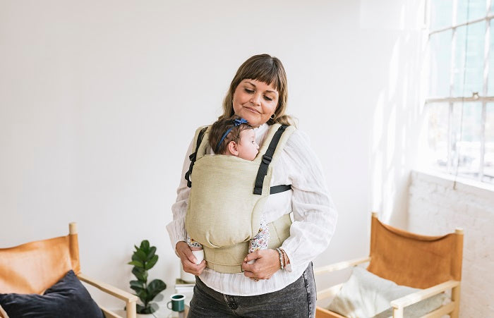 A smiling mother carrying her baby in an ergonomic Free-to-Grow Baby Carrier Linen Moss from Tula.