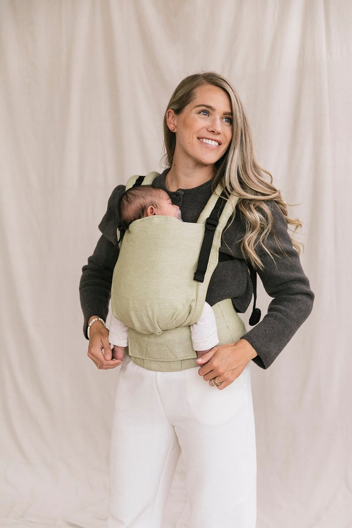 A smiling mother carrying her baby in an ergonomic Free-to-Grow Baby Carrier Linen Moss from Tula.