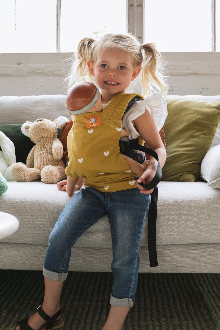 A little girl wearing the Tula Mini Toy and Doll Carrier Play.