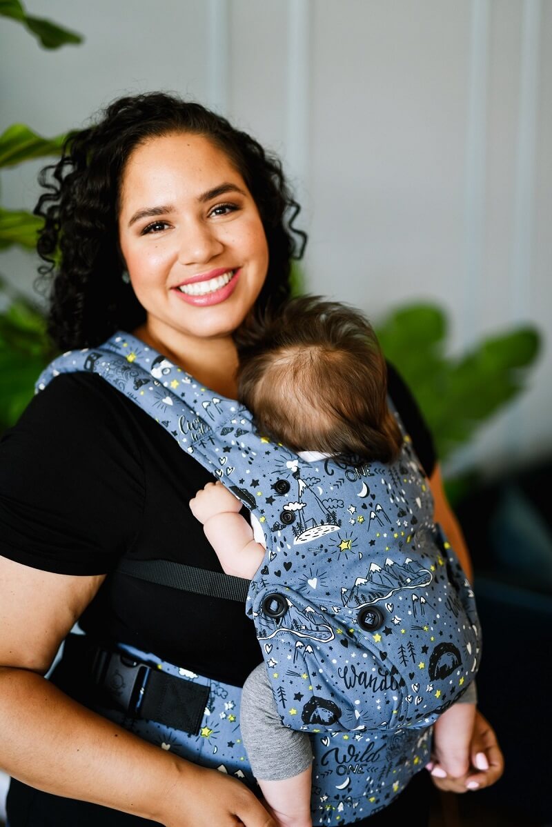 A caregiver smiling into the camera while her baby is sleeping in the Wander Explore Baby Carrier.