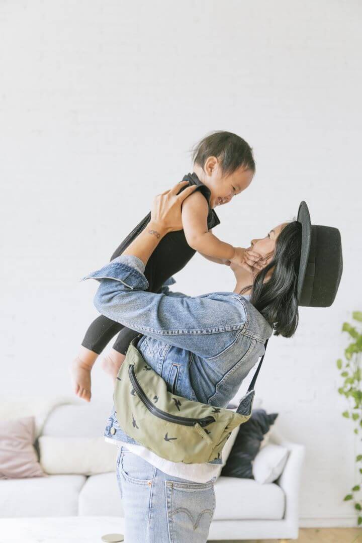 Caregiver playing with toddler wearing a compact travel baby carrier in its pouch over their shoulder.